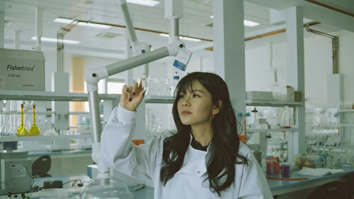 A woman in a lab coat and blue gloves carefully working with a pipette, holding two test tubes in her hands. She is in a laboratory setting, with a rack of test tubes visible in the foreground and a white wall in the background. The image is focused on her hands, emphasizing the scientific process.